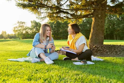 Outdoor meeting of teenage girl and woman of psychologist social worker, women talking sitting on green lawn in park, comfortable environment for talking with young people, mental help consultation