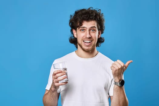 handsome bearded man in white t-shirt with drinking glass of water pointing to the side on blue background