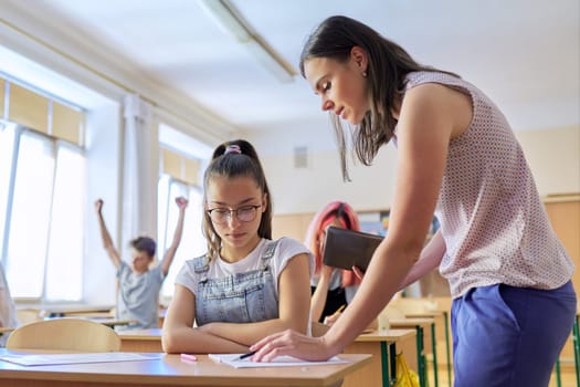 Young woman teacher teaches lesson in class of teenage children, teacher stands near school desk with girl student, checks knowledge. Education, school, college, teaching concept