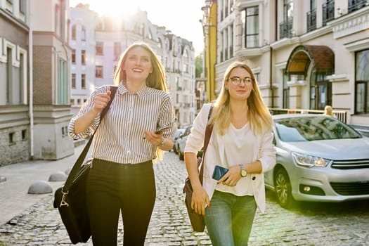 Two young beautiful happy women university students walking along city street, women smiling laughing looking forward