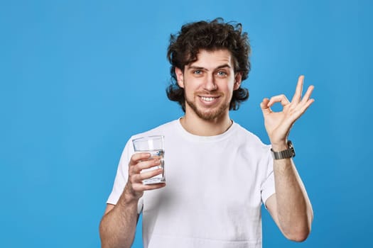 Young smiling man in white t-shirt with drinking glass of water showing Ok gesture on blue background
