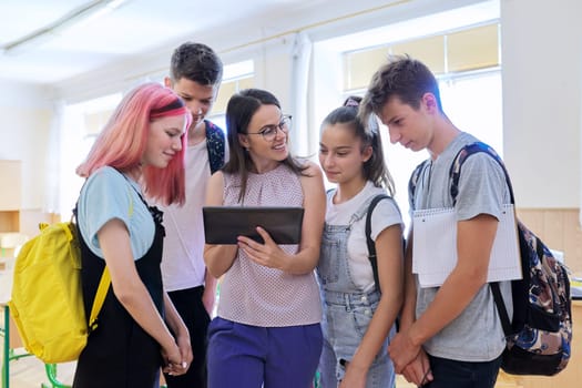 Group of teenage students and young female teacher with digital tablet in classroom, children and tutor looking at computer screen. Education, school, college, schoolchildren concept