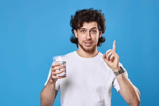 Young curly man in white t-shirt with drinking glass of water pointing up on blue background