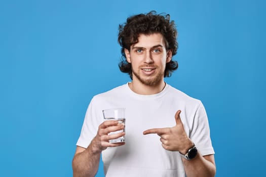 young positive man in white t-shirt showing drinking glass of water on blue background