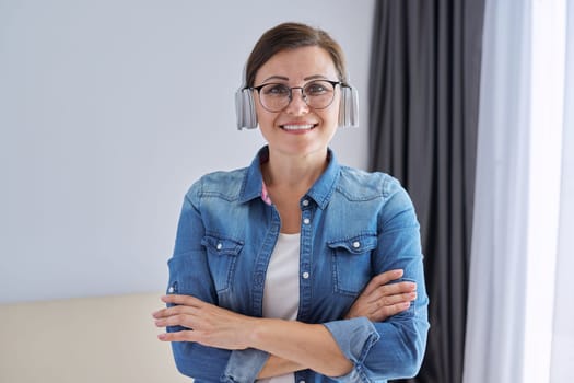 Portrait of confident middle aged mature woman wearing glasses headphones with folded hands, in home interior. Business woman, freelancer, teacher, psychologist, counselor, blogger working remotely
