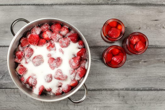 Tabletop view - large steel pot with strawberries covered in crystal sugar, three pickled strawberry glass bottles next to it. Homemade compote preparation.