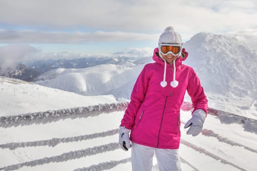 Young woman in pink jacket, wearing ski goggles, leaning on snow covered fence, smiling, white mountains in background.