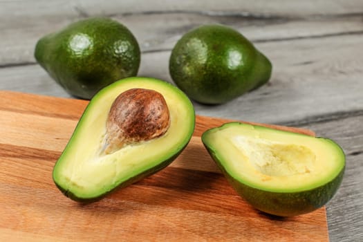 Avocado cut in half, seed visible, on wooden chopping board, two more whole pears in background.