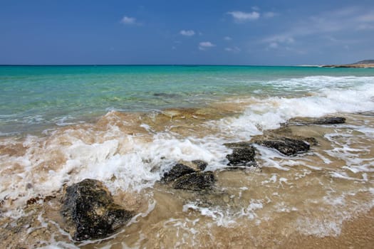 Small sea waves crushing on black rocks at sandy beach, beautiful blue crystal clear water in background. Karpass, Northern Cyprus