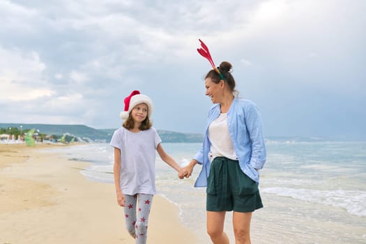 Happy mom and daughter child in Santa hat walking holding hands along the beach. Family celebrating Christmas and New Year at tropical resort, vacations travel tourism concept
