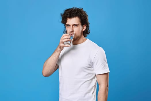 handsome curly man drinks water from a glass on blue background