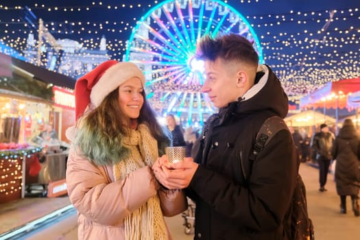 Happy teenagers couple boy and girl in Santa hat at Christmas market warming hands with candle. Lights, bright garlands of evening city, ferris wheel background