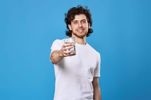 happy handsome man in a white t-shirt showing drinking glass of water on blue background. focus on glass