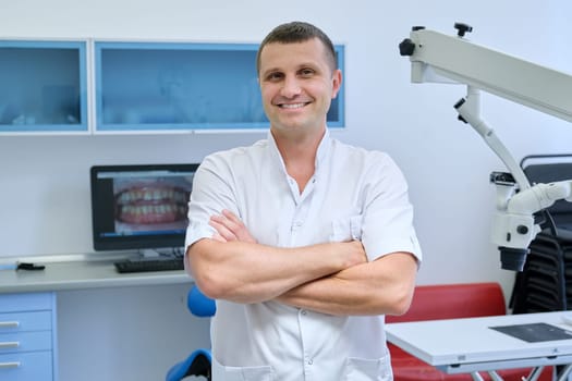 Portrait of confident friendly male dentist with crossed arms, smiling professional doctor looking at camera in dental clinic