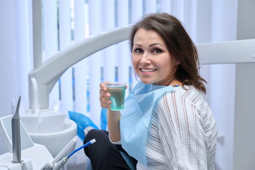 Happy mid adult woman patient in dental chair with glass of water, smile with healthy white teeth. Dental treatment, medicine dentistry and health care concept