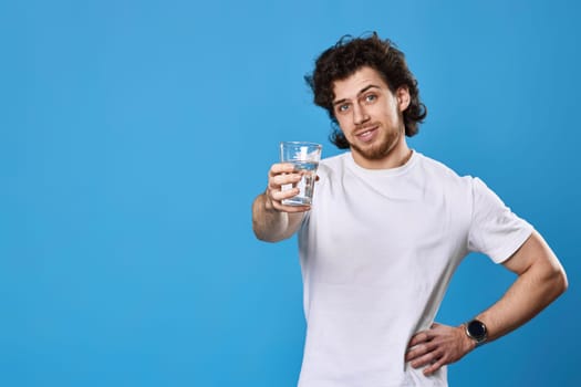Young happy curly man showing drinking glass of water on blue background. focus on glass