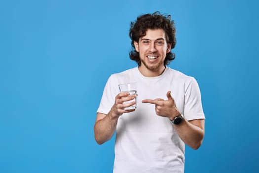 happy handsome man in white t-shirt showing drinking glass of water on blue background