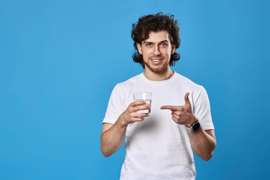 curly handsome man in white t-shirt showing drinking glass of water on blue background
