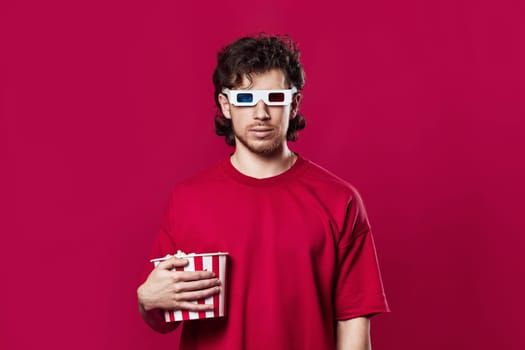 handsome bearded man in 3d glasses holding big bucket of popcorns on magenta background