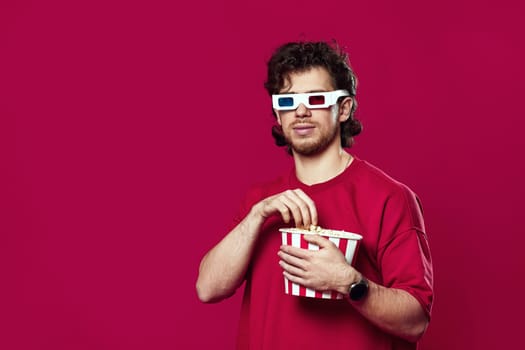 handsome bearded man in 3d glasses holding big bucket of popcorns on magenta background