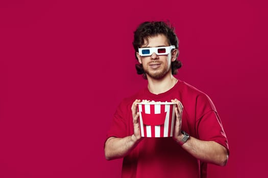 handsome bearded man in 3d glasses holding big bucket of popcorns on magenta background
