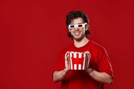 handsome bearded man in 3d glasses holding big bucket of popcorns on red background