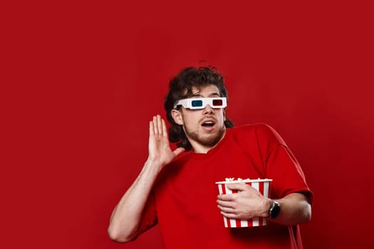 scared handsome man in 3d-glasses holding bucket of popcorn on red background