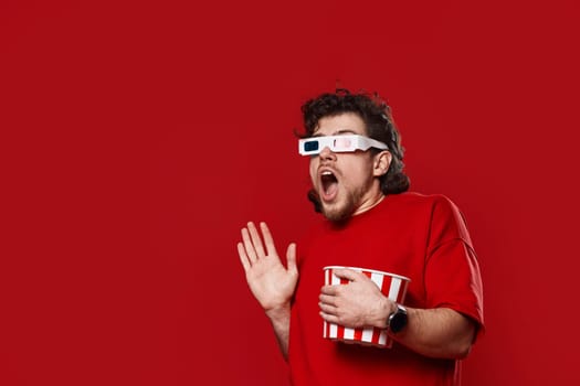 Shocked scared handsome man in 3d-glasses watching movie film hold bucket of popcorn on red background. copy space