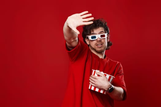 scared handsome man in 3d-glasses holding bucket of popcorn on red background