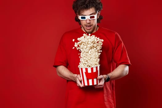 young handsome man in 3d glasses, red t-shirt jumping with big bucket of popcorn on red background