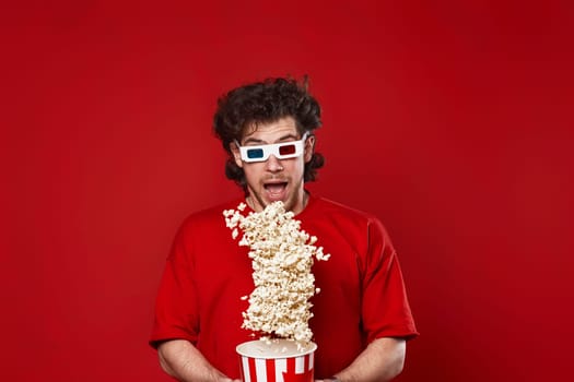 young handsome man in 3d glasses, red t-shirt jumping with big bucket of popcorn on red background