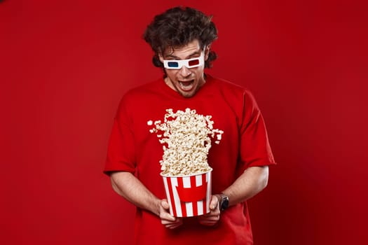 young handsome man in 3d glasses, red t-shirt jumping with big bucket of popcorn on red background