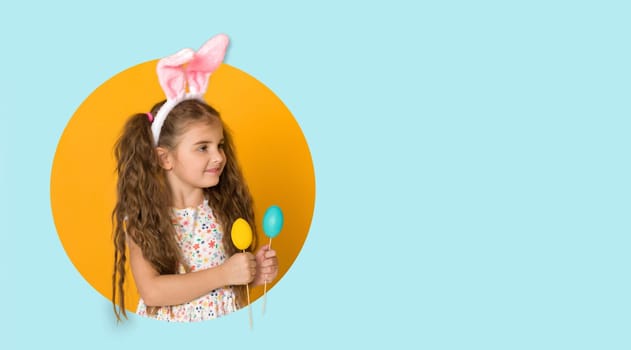 smiling curly caucasian little child girl with Easter bunny ears and colorful eggs on yellow and blue background. copy space