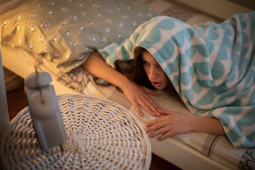 A frightened young woman hides under a blanket from the sound of her alarm clock. A girl under a soft blanket.