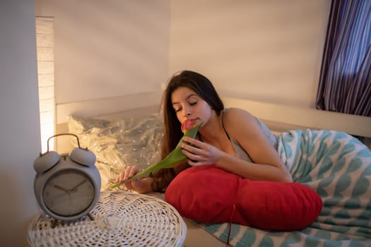 teenage girl holds a tulip in her hand and sniffs it. A large alarm clock stands on the bedside table.