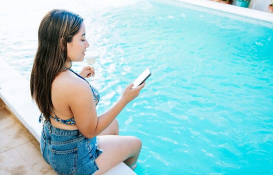 Girl sitting on the edge of the swimming pool holding drink and checking cell phone. Smiling girl on vacation in swimming pool checking cell phone and holding cocktail