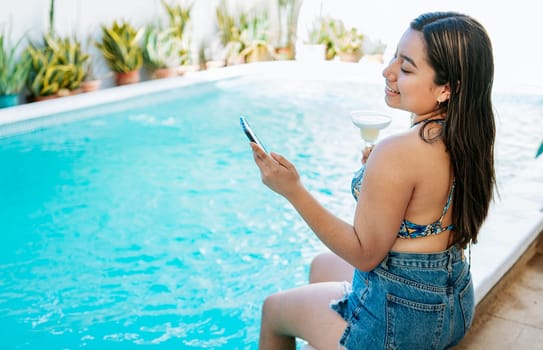 Smiling girl on vacation in swimming pool checking cell phone and holding cocktail. Girl sitting on the edge of the swimming pool holding drink and checking cell phone