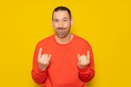 Hispanic man with a beard wearing a red pullover making the rock gesture with arms close to the body isolated on yellow studio background, isolated on yellow background.