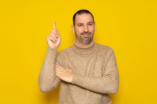 Bearded hispanic man wearing a beige turtleneck raising his index finger asking for a turn to express the great idea he had, he feels proud for having found a solution to the problem