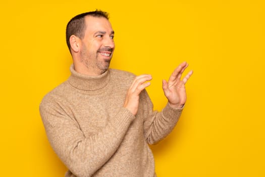Bearded hispanic man in a turtleneck ducking to the side in embarrassment, tries to push away from something or someone with his hands. Isolated on yellow background