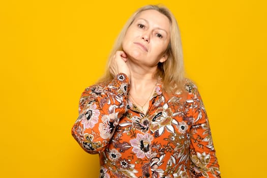 Caucasian blonde woman in a patterned dress scratching her neck with a carefree attitude while hesitating to choose one thing or the other, isolated over yellow background