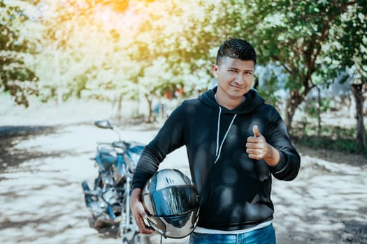 Portrait of motorcyclist holding safety helmet with thumb up. Smiling motorcyclist man holding safety helmet on the road. Motorcyclist giving thumbs up holding safety helmet on the road