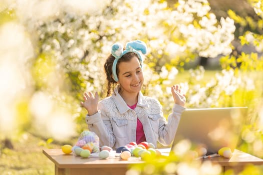 happy teen girl wear bunny ears paints Easter eggs with a laptop in the garden.