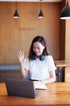 Portrait of a teenage Asian woman using a computer and notebook to study online via video conferencing on a wooden desk in library.