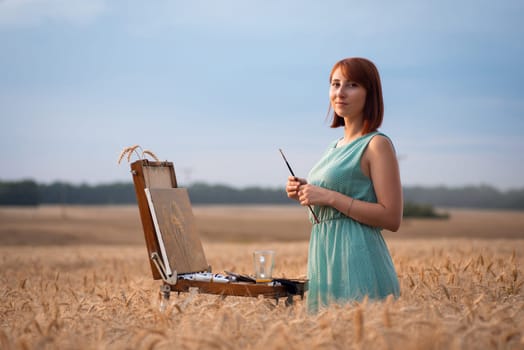 Beautiful young girl painter near easel with unfinished work looking to the camera.