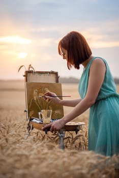 Girl artist painting ears of wheat in field of rye at sunset