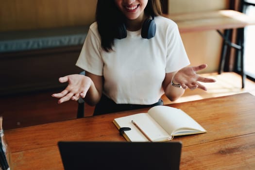 Portrait of a teenage Asian woman using a computer and notebook to study online via video conferencing on a wooden desk in library.