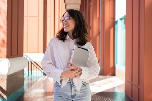 Businesswoman in glasses looking away, smiling and holding digital tablet, standing outdoor. Female freelancer holding tablet while walk outside