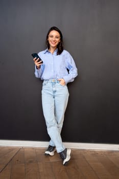 Happy hipster girl wear glasses, types text message on mobile phone, enjoys online communication, types feedback on black studio background. Full length, looking at camera and smile