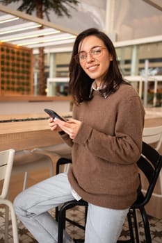 Portrait of positive cheerful girl use smart phone look at camera share social media news wear stylish trendy jumper sitting bar stool. Female smartphone user. Woman in glasses holding smartphone